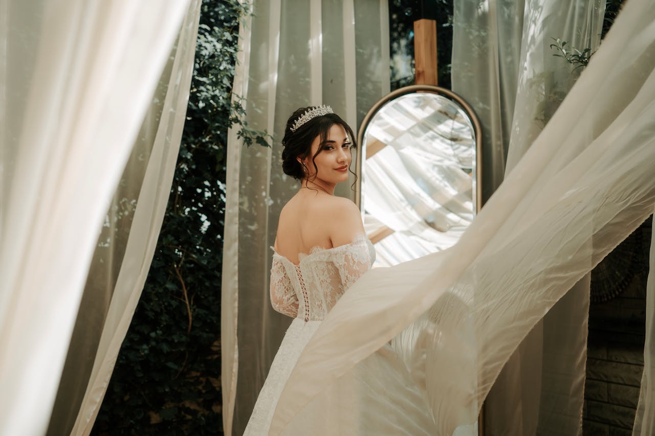 A beautiful bride in a lace wedding dress looks back towards a mirror surrounded by flowing curtains.