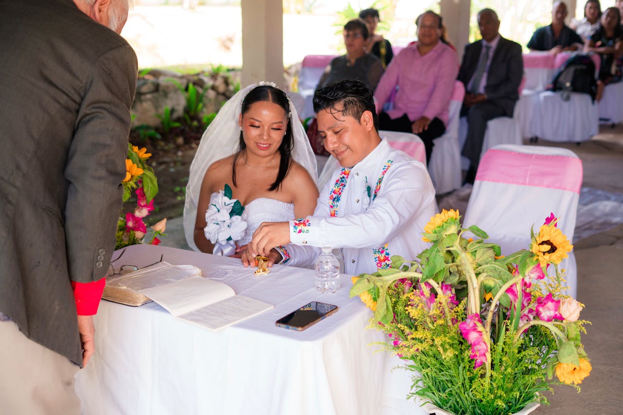 Bride and groom sign wedding documents during an outdoor ceremony with colorful floral decorations.